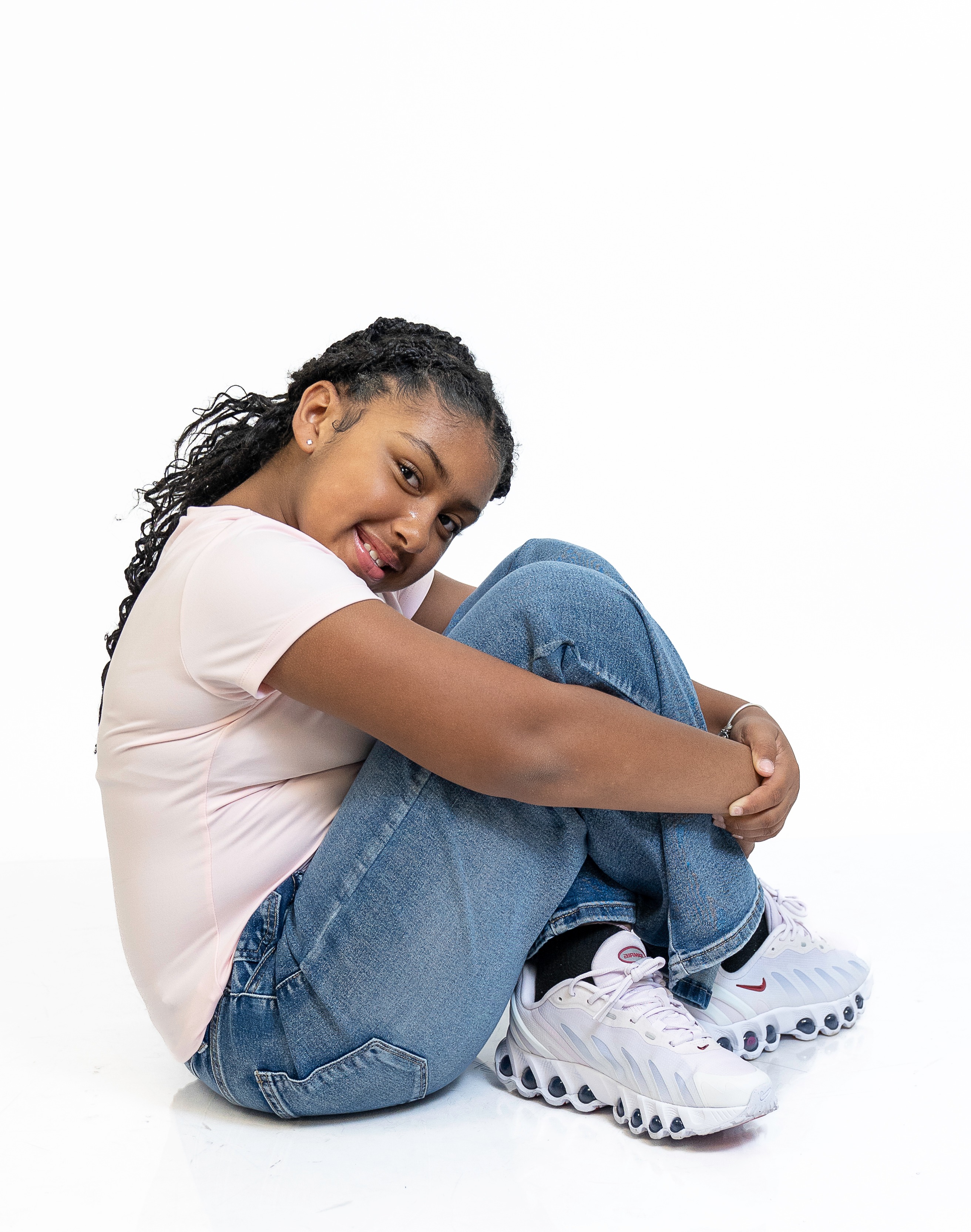 Eleaya Neveah full-body photo, seated on the floor hugging her knees, pink tee and jeans